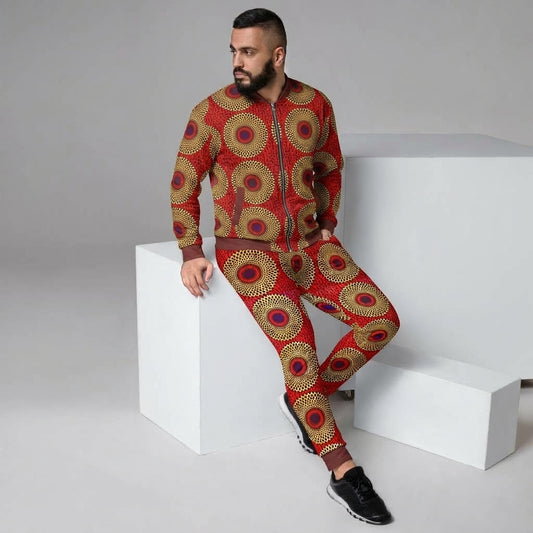 A man wearing a matching red zip-up jacket and pants set with a gold and brown circular African wax print pattern, posing on white geometric blocks against a gray studio background.