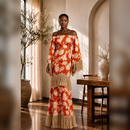 A woman stands indoors wearing an off-the-shoulder maxi dress with a bold orange and cream floral pattern and long fringe trim at the hem and sleeve cuffs. The setting is a warm, sunlit room with beige walls, an arched doorway, a wooden table, and potted plants.