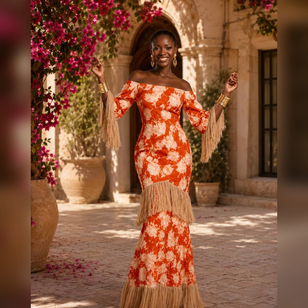 A woman stands outdoors in a sunlit courtyard wearing an off-the-shoulder orange and cream floral mermaid gown with long fringe sleeves and a matching fringe hem. Pink flowers and stone architecture are visible in the background.