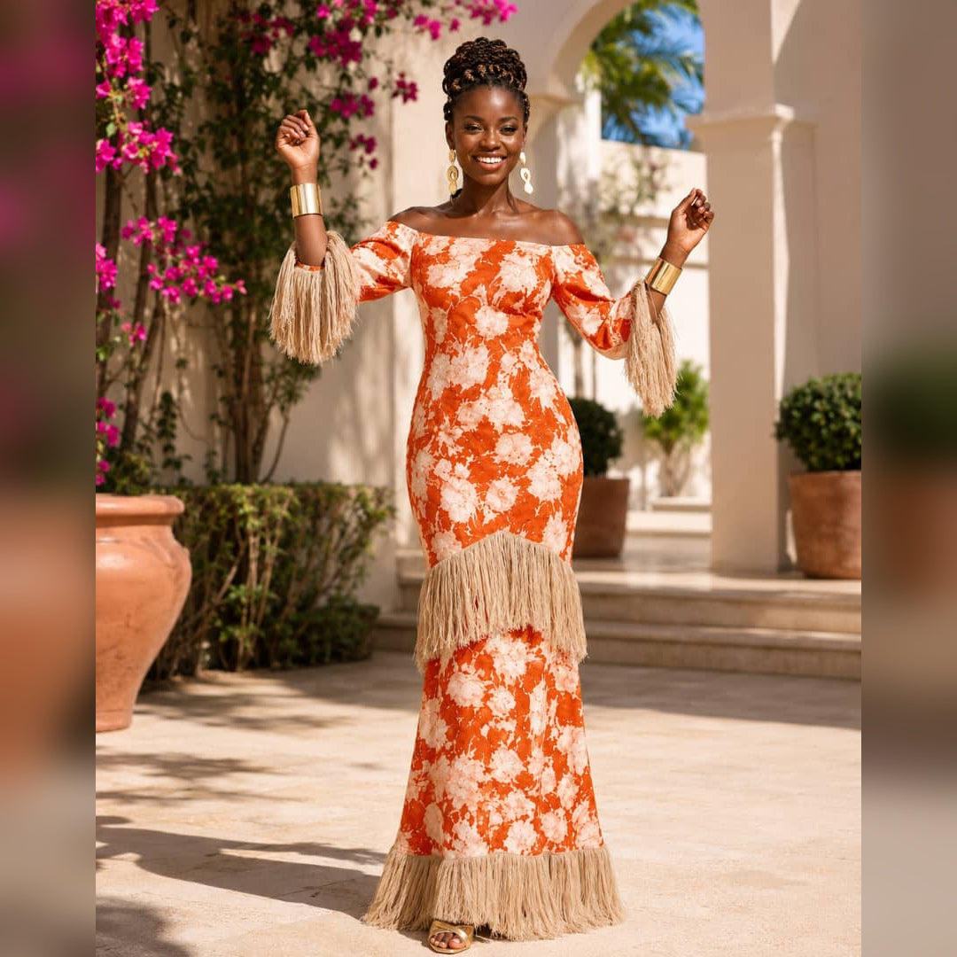 A woman stands outdoors in a sunlit courtyard wearing an off-the-shoulder orange and cream floral maxi dress with beige fringe detailing on the sleeves, waist, and hem. The background features white columns, potted plants, and bright pink flowers.