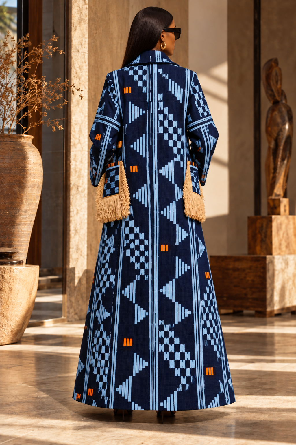 A woman viewed from behind wearing a long navy maxi dress with a light blue geometric pattern and small orange accents, standing in a sunlit indoor setting with large vases and sculptures.