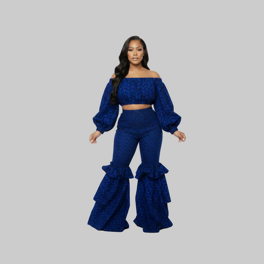 A woman modeling a matching royal blue off-the-shoulder crop top and high-waisted flare pants set with a textured pattern and layered ruffle hems, set against a solid gray background.