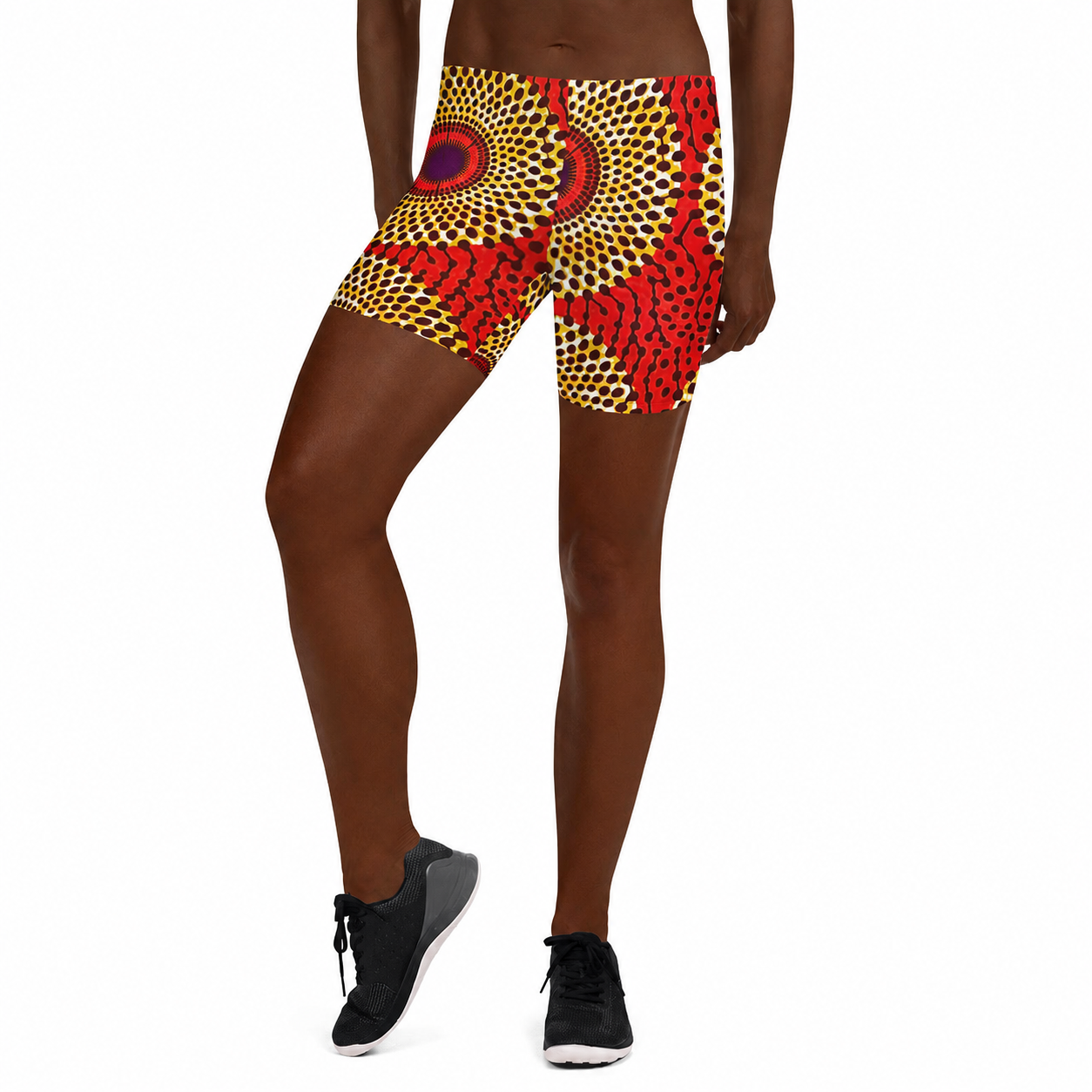 A model wearing high-waisted athletic shorts with a vibrant red, yellow, and black African wax print pattern, paired with black athletic shoes, shown against a plain white background.