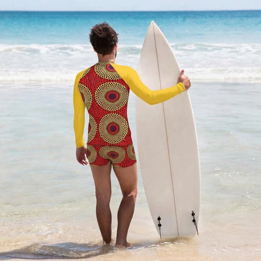 A man on a beach stands in shallow water holding a white surfboard, wearing a one-piece swimsuit with bright yellow sleeves and a red body featuring a circular geometric pattern in yellow, black, and white.