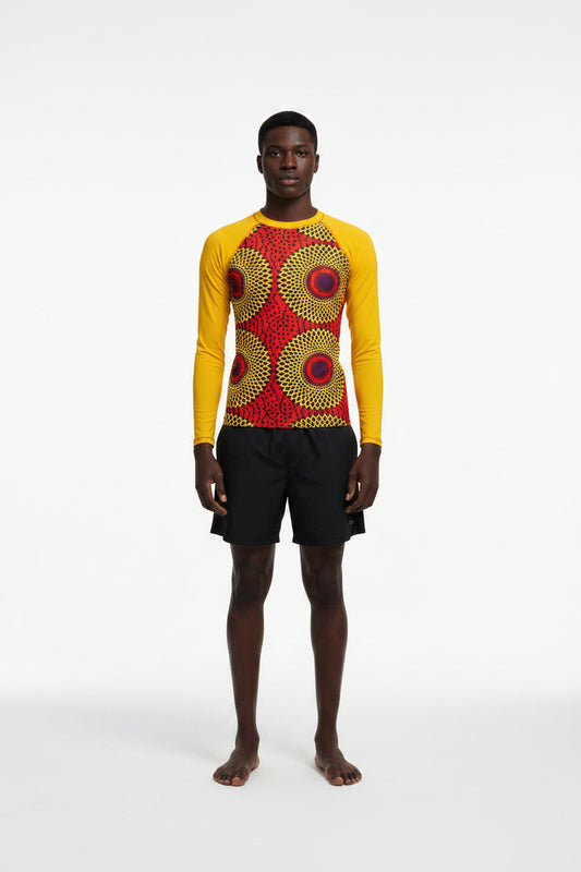 A man standing at the beach wearing a yellow rash guard with a Rekiana African print design, including a sunglasses and shorts.