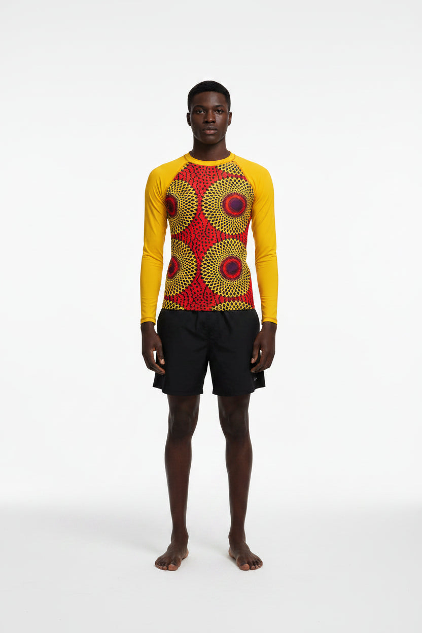 A man standing at the beach wearing a yellow rash guard with a Rekiana African print design, including a sunglasses and shorts.
