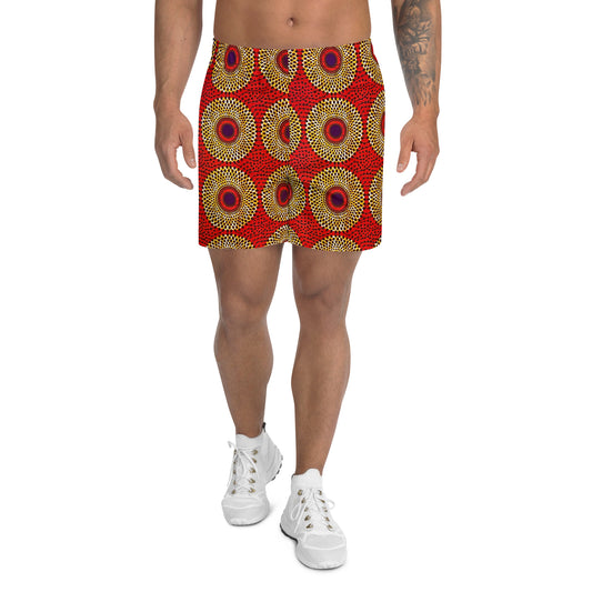 A man wearing knee-length Rekiana African print athletic shorts in a red, yellow, and black geometric pattern, featuring a flat white drawstring elastic waistband and mesh side pockets.