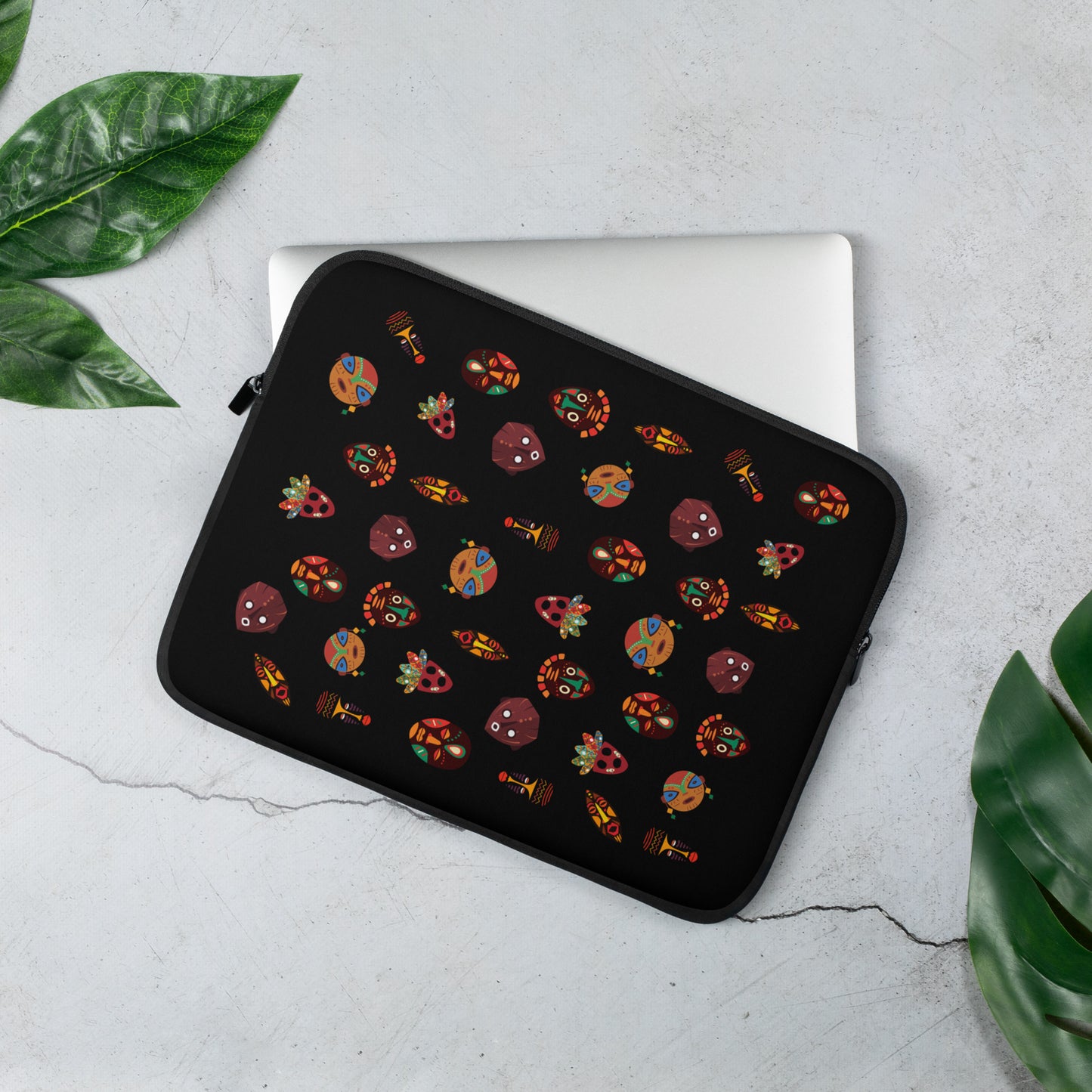 A 13 inch laptop sleeve with vibrant, multicolored tribal masks, placed on a flat surface with a laptop and a green plant in the background.