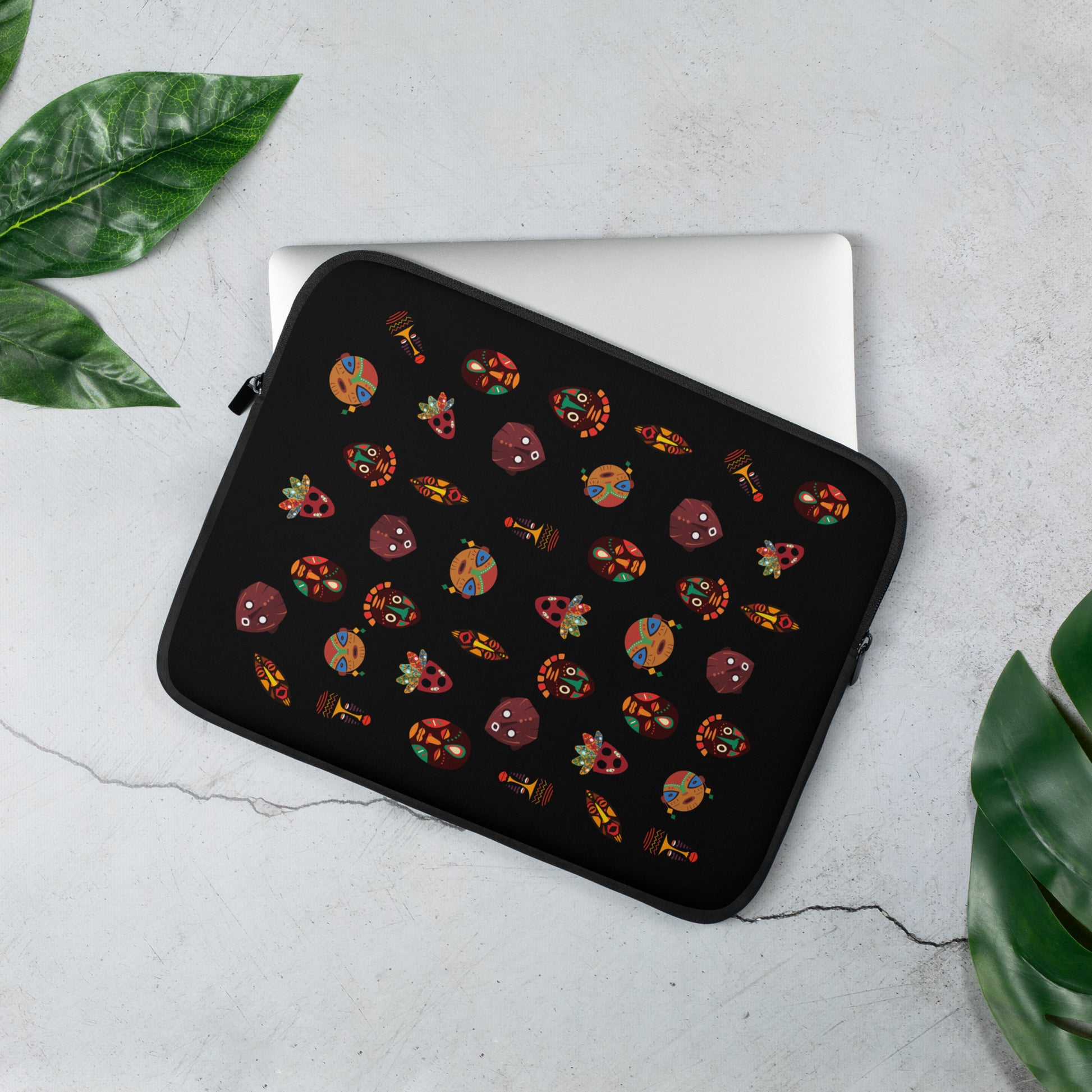A 13 inch laptop sleeve with vibrant, multicolored tribal masks, placed on a flat surface with a laptop and a green plant in the background.