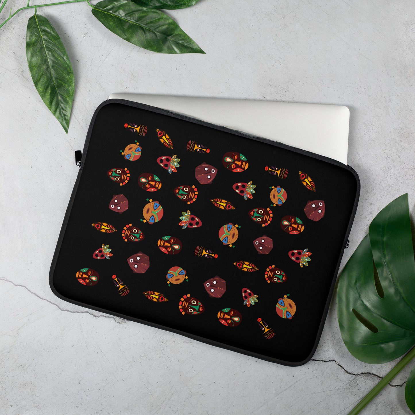 A 15 inch laptop sleeve with vibrant, multicolored tribal masks, placed on a flat surface with a laptop and a green plant in the background.