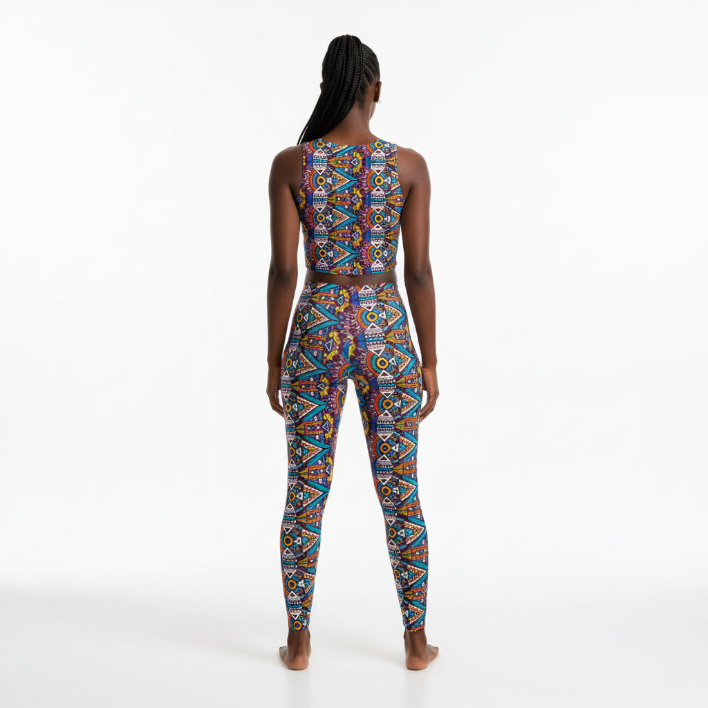 A woman stands with her back to the camera wearing a sleeveless, high-waisted one-piece jumpsuit with a vibrant geometric pattern in blue, red, orange, yellow, and white on a plain white studio background.
